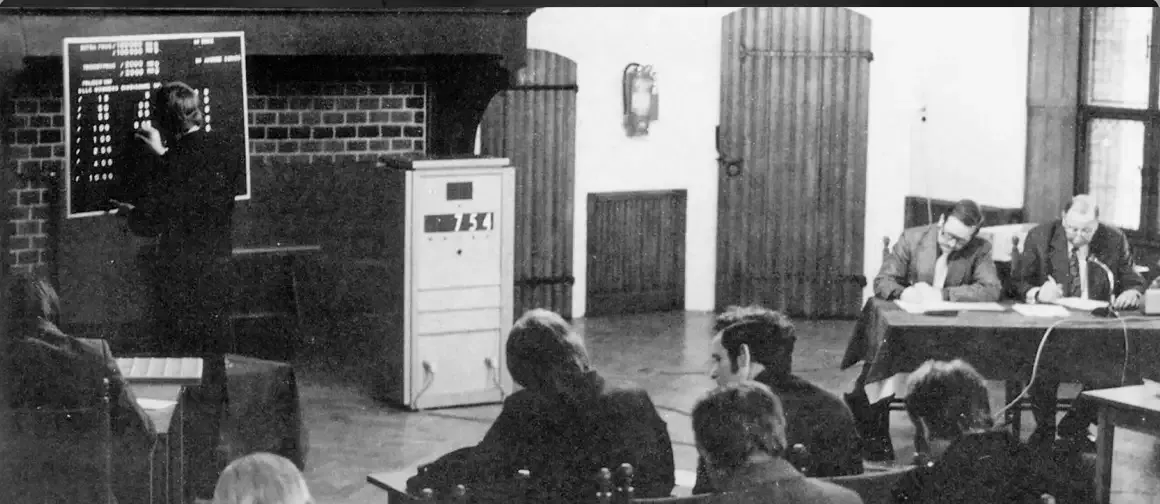 Historische afbeelding van een trekking met deelnemers en een presentator aan een bord.