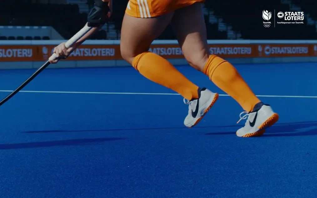Close-up van een speler in oranje sportkleding die over een blauwe hockeybaan rent met een hockeystick.