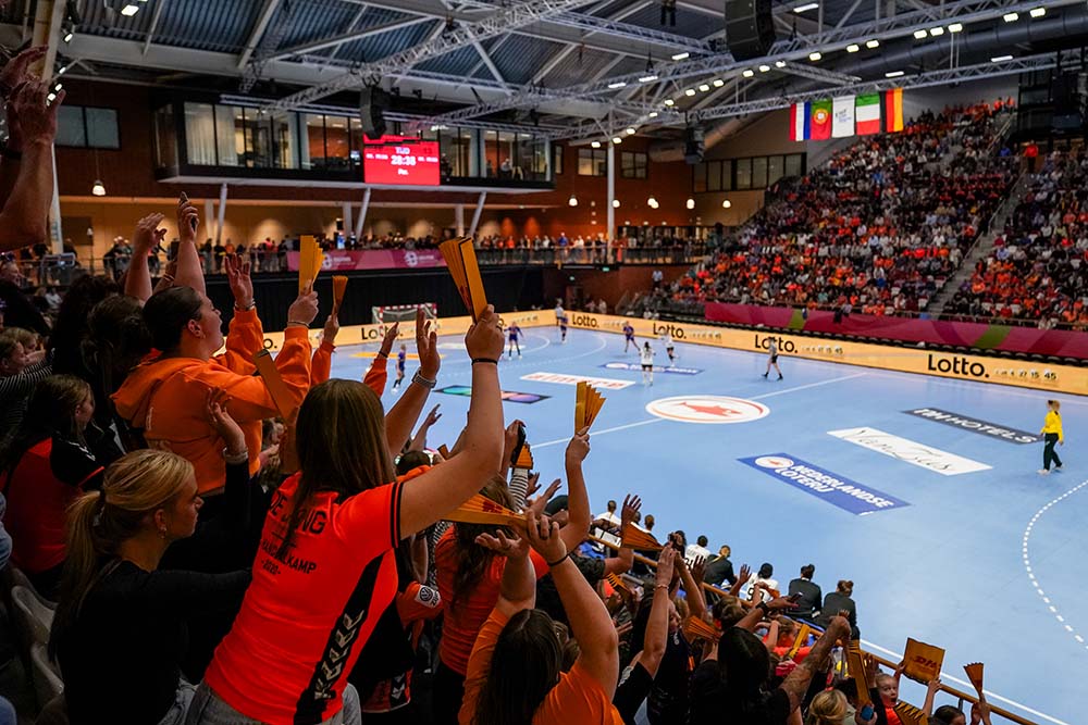 Spectaculaire handbalmatch met enthousiaste supporters in oranje shirts die gouden kaarten omhoog houden.
