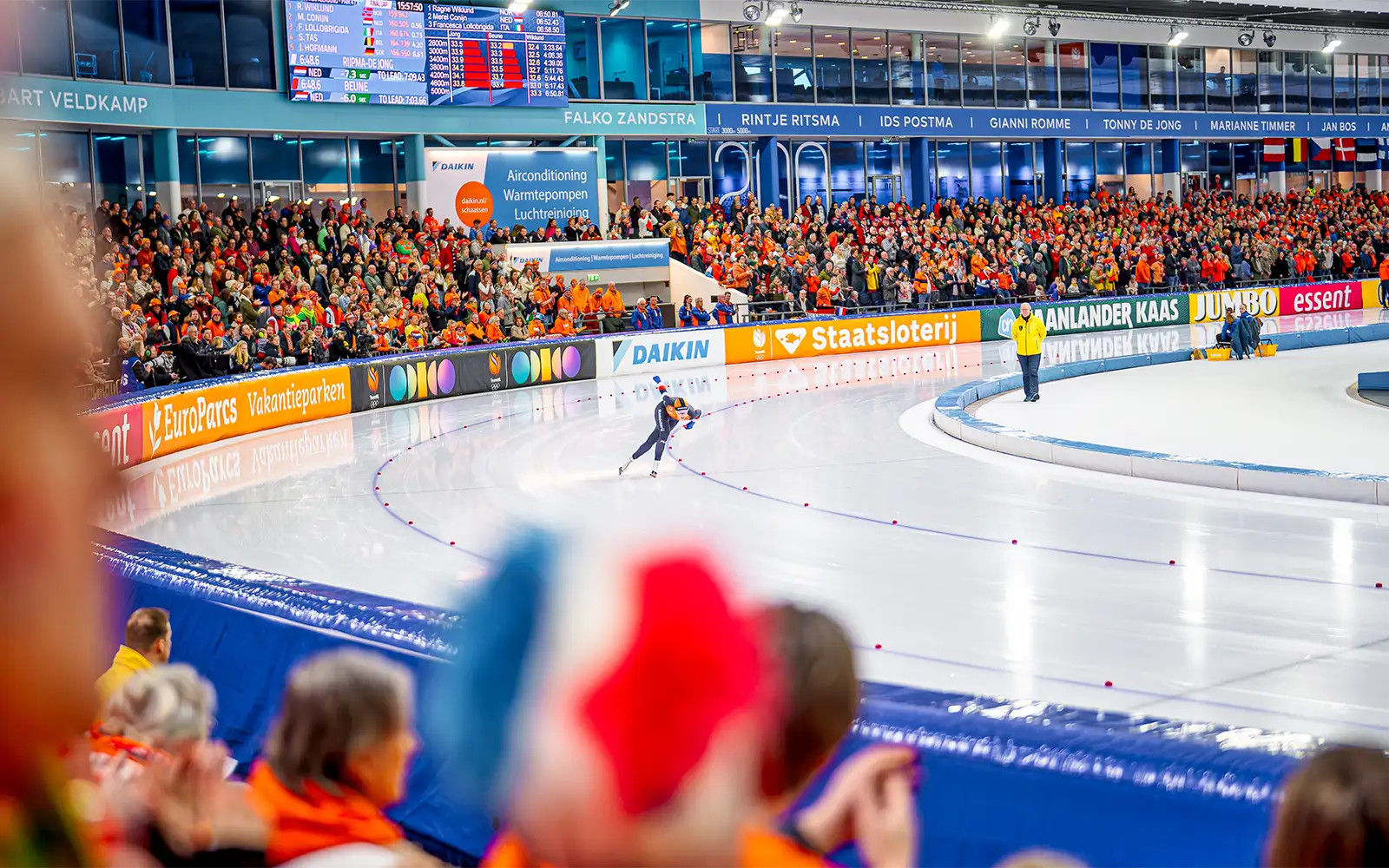Schaatser in actie met enthousiaste menigte in oranje kleding op tribunes bij een evenement.