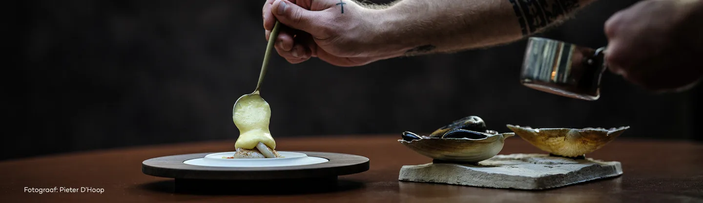 Een hand leegt een lepel met een saus op een wit bord, met losse schelpen op een steen.